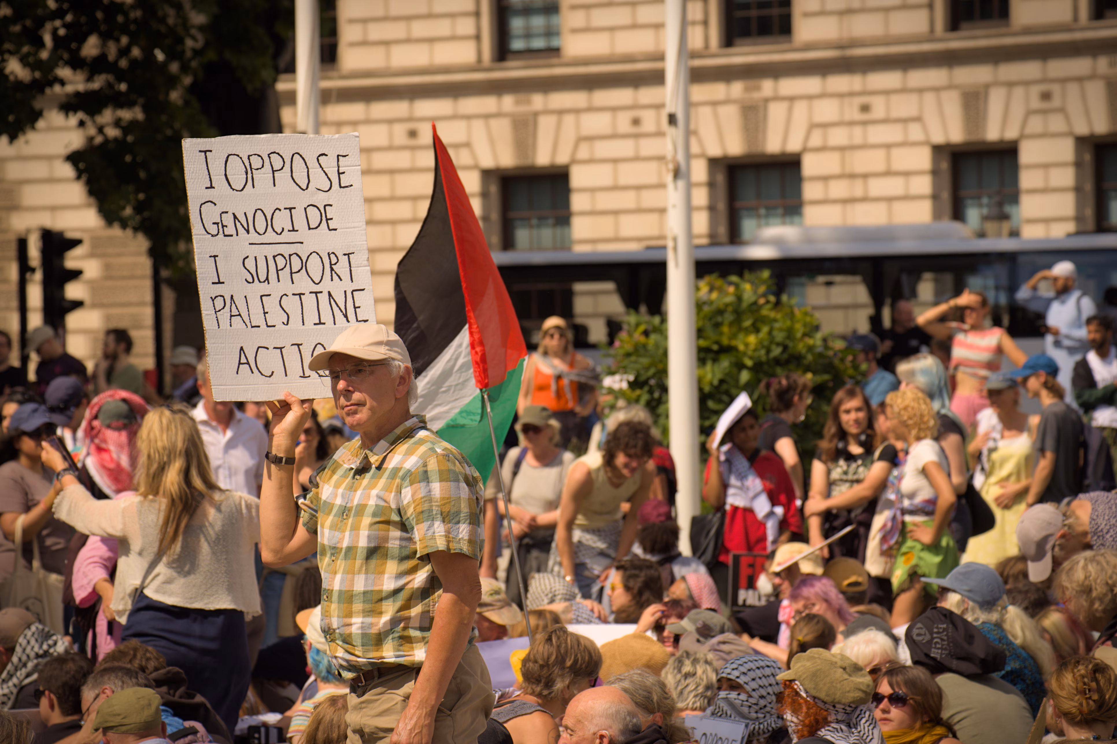 An elderly man dressed in a casual short-sleeved shirt and chinos wearing a tan cap holding up a sign reading, "I oppose genocide; I support Palestine Action," in front of a crowd of protestors and a Palestinian flag.
