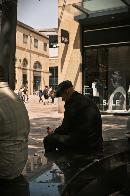 A street photograph of a man sat on a public bench in the summer, wearing a flat cap, checking his phone.